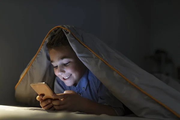 Little boy in bed under a blanket looking at the smartphone at night. Teen boy lying on front in the bed and using smartphone. 7 years old child boy using smartphone at night in bed