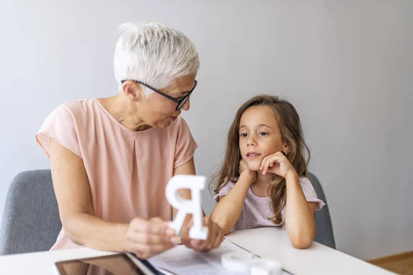 Girl visiting speech therapist. Mature female therapist helping girl in speech therapy exercise. Cute little girl at speech therapist office. Mature woman teacher and little girl on private lesson