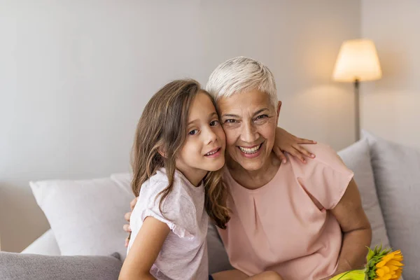 Granddaughter embracing her grandmother in living room. Lovely little girl with her grandmother looking at the camera. Girl and grandma on a sofa Smiling senior woman and girl embracing