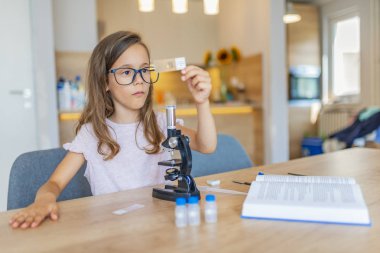 Kendine güvenen küçük kız evde mikroskop kullanır. Güzel ilkokul öğrencisi bilim projesi için bir mikroskop kullanır. Mikroskop kullanırken küçük kız konsantre