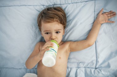 Toddler boy drinking milk from a bottle on blanket. Baby boy feeding with bottle of milk. Time to sleep. Baby drinking milk while lying on bed. Time to feeding.  clipart