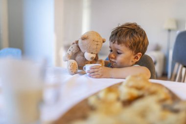 Çocuk kahvaltı yapıyor. Çocuk oyuncak ayıyı besliyor, süt içiyor ve meyveli mısır gevreği yiyor. Pencerenin önündeki mutfakta beyaz yemek masasında oturan küçük çocuk. Çocuklar yer. Genç çocuklar için sağlıklı beslenme.