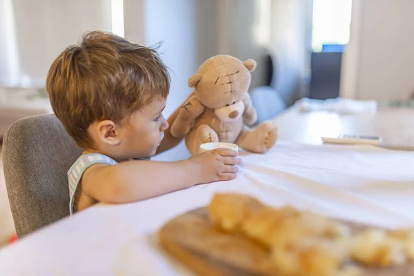 かわいい幼児の男の子は キッチンで椅子に座って テディベアのおもちゃで遊んで ヨーグルトを飲みます 子供はテディベアのおもちゃを供給します 朝食を食べる子供 ミルクの子供 — ストック写真