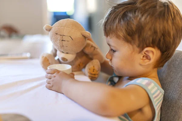 Cute little boy giving glass of milk to teddy bear. Playing with teddy bear at home. Childhood happiness concept. A boy is sitting and eating with Teddy bear. Child having breakfast. Kid feeding teddy bear toy