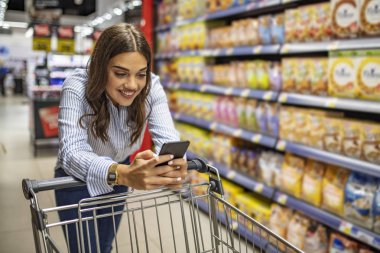 Alışveriş arabasına yaslanan, cep telefonu kullanan ve gülümseyen bir kız görüntüsü. Kadın market arabasıyla ürün satın alıyor. Kadın süpermarkette alışveriş yapıyor.