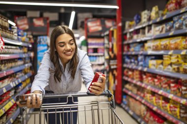 Süpermarket koridorunda alışveriş arabası olan bir kadın. Marketten yiyecek almak. Market alışverişi. Marketten alışveriş yapan genç ve güzel bir kadın. 