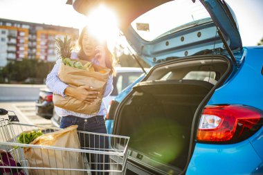 Kadın, marketten alışveriş yaptıktan sonra arabaya biniyor. Alışverişi getiriyorum. Market alışverişi yapan mutlu kız. Alışveriş yaptıktan sonra arabaya çanta koyan kadın.