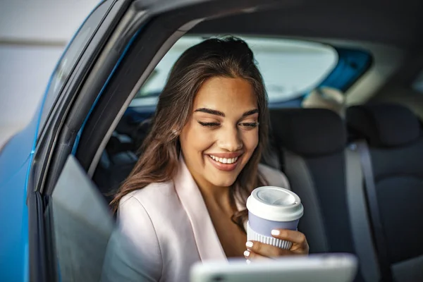 Young, beautiful woman sitting in the back seat of the car with a tablet in hand and drinking coffee. Female enterpreneur with coffee watching podcast on tablet PC