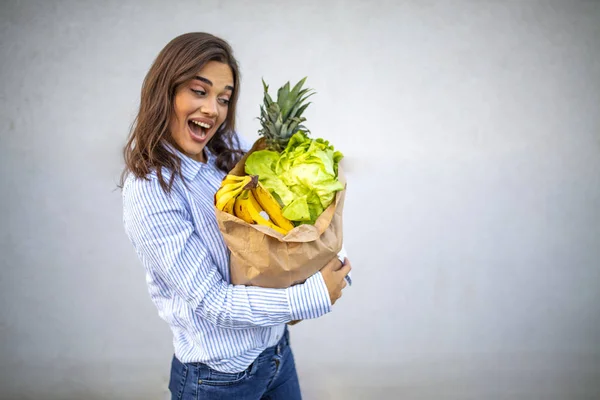 Young woman holding paper bag of groceries from supermarket with a happy face standing and smiling with a confident smile showing teeth. Beautiful smiling woman holding paper shopping bag full of vegetables and groceries