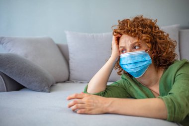 Tıbbi maskeli genç bir kadın kendini karantinaya almak için evde karantinada kalıyor. Ev karantinası, COVID-19 önleme, Coronavirüs salgını durumu. Evde koruyucu maskeli bir kadın var..