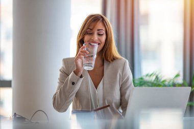 Bir dizüstü bilgisayarla ve ofiste içme suyuyla çalışan mutlu girişimci. Ofis çalışanı bir bardak suya uzanıyor. Gülümseyen iş kadını ofisteki masasında bir bardak su içiyor, mola veriyor..