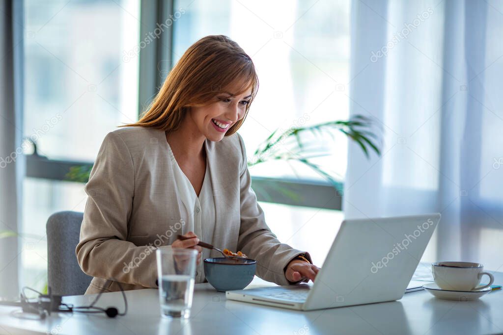Beautiful young woman sitting at the desk in an office and eating lunch, using laptop at the same time. Business woman eating lunch at her workplace looking at the laptop screen.