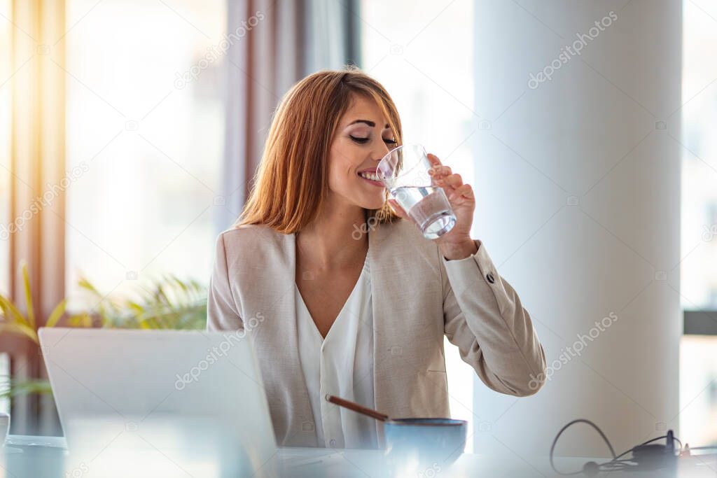 Businesswoman drinking a glass of water at her desk in the office. Young woman working in her offfice. Woman drinking water from glass in the office in the morning with sunlight