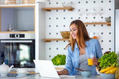 Mutfakta dizüstü bilgisayar kullanan güzel bir kadın. Genç bir kadın mutfak tezgahına eğilip dizüstü bilgisayara bakıyor. Güzel kadın kahvaltı yapıyor ve dizüstü bilgisayar kullanıyor.. 