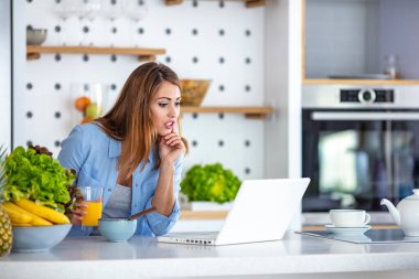Modern sabah rutinleri. Evdeki mutfakta kahvaltı ederken dizüstü bilgisayar kullanan genç bir kadının fotoğrafı. Genç, neşeli bir kadın dizüstü bilgisayar kullanıyor ve mutfakta mısır gevreği yiyor.