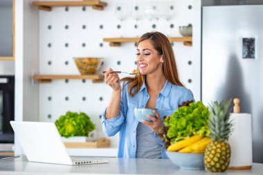 Güzel kız evde mutfakta dururken dizüstü bilgisayar kullanıyor ve gülümsüyor. Genç bir kadın dizüstü bilgisayar kullanıyor ve mutfakta kahvaltı yapıyor..