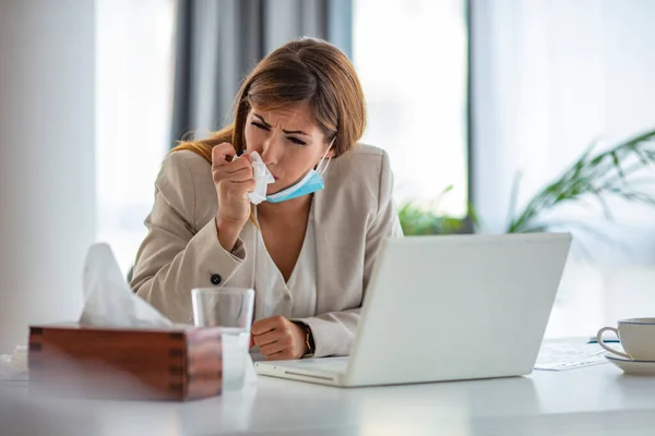 Shot of a young businesswoman blowing her nose while working in an office. Women are sneezing and are cold. She is in the office. I hope this flu goes away quickly.