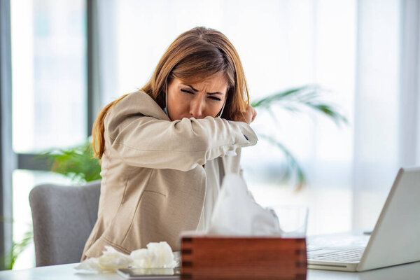 Young businesswoman coughing into elbow in the office. Cropped shot of a businesswoman working in her office while suffering from allergies. Women are sneezing and are cold. She is in the office.