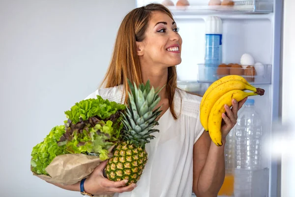 Woman picking up some fruits and veggies from the fridge. Closeup of a cheerful young woman picking some fruit and veggies from the fridge to make some healthy breakfast.