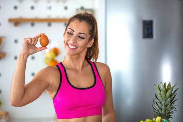 A fitness woman is showing off chicken eggs. She stands with them in her hands and a kitchen in the background. Smiling Woman Holding Egg. Fitness woman prepare healthy breakfast.