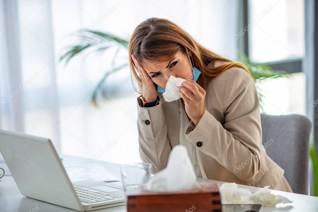 Shot of a young businesswoman blowing her nose while working in an office. Women are sneezing and are cold. She is in the office. I hope this flu goes away quickly. 