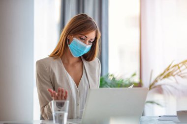 Ofiste dizüstü bilgisayarla çalışan koruyucu maskeli bir iş kadını. Ofiste iş raporları üzerinde çalışan yüz maskeli bir kadın girişimci. Boşluğu kopyala.