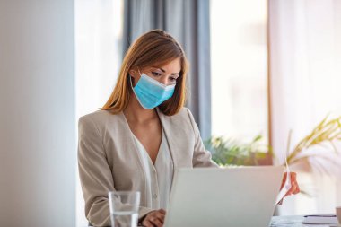 Ofiste dizüstü bilgisayarla çalışan koruyucu maskeli bir iş kadını. Ofiste iş raporları üzerinde çalışan yüz maskeli bir kadın girişimci. Boşluğu kopyala.