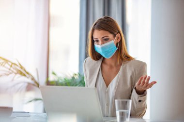Ofiste dizüstü bilgisayarla çalışan maskeli bir iş kadını. Kavram, bulaşıcı hastalık, koronavirüs. Ofisteki genç kadın, koronavirüsün yayılmasını önlemek için koruyucu maske takıyor.