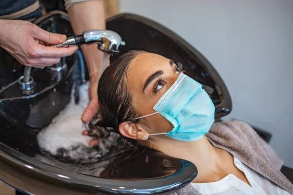 Happy woman wearing face mask while hairdresser is washing her hair at the salon during COVID-19 epidemic. Young woman with surgical mask on face at hairdresser salon. Covid-19 concept.