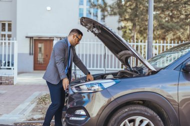 Takım elbiseli profesyonel bir adam kaputu açık bir şekilde arabasının yanında duruyor, güneşli bir caddedeki motoru inceliyor..