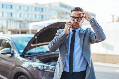 Arızalı arabasının yanında duran profesyonel bir adam yardım için telefon ediyor. Açık araba kaputu ani bir soruna işaret ediyor ve adamın davranışı aciliyet ve endişeyi vurguluyor..