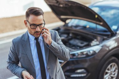 Profesyonel giyimli bir adam yol kenarında duran arabasının yanında bir telefon görüşmesi yapar. İş ortamında acil durum ve sorun çözme işlemleri yapar..