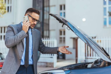 Takım elbiseli bir işadamı kaputu açık bir arabanın yanında duruyor, yol yardımı için bir telefon görüşmesi yapıyor, mekanik sorunlar veya şehir ortamında meydana gelen bir arıza hakkında endişeli görünüyor..