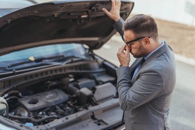 Profesyonel giyimli bir adam bir araba arızasını açık bir kaputla inceliyor ve hayal kırıklığı gösteriyor. Açık hava ortamında iş günlerinde beklenmedik araç sorunları vurgulanıyor.