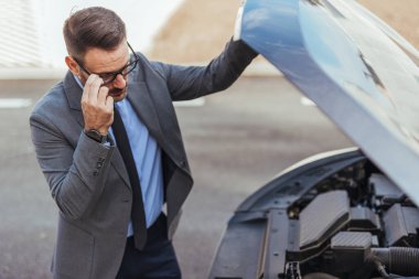 Takım elbiseli profesyonel bir adam kaputun altındaki araba motorunu inceleyerek potansiyel bir sorunu analiz eder. Sahne, otomotiv bakımının zorluklarıyla ilgili endişe, sorumluluk ve sorun yaratmayı betimliyor.
