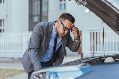 Takım elbiseli bir iş adamı açık kaputu inceliyor, açık şehir ortamında olası bir arıza hakkında stresli görünüyor..