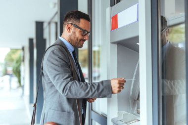 Profesyonel bir adam açık hava ATM 'sinden banka hizmetlerine erişiyor, kart işlemlerini güvenli bir şekilde yürütüyor. Modern finansal teknolojiyi ve iş kolaylığını temsil ediyorum..