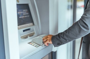Bankamatik işleten gri ceketli bir adam, modern bir binadan para çekmek gibi bankacılık işleriyle uğraşıyor..