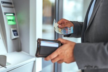 Takım elbiseli bir adam, modern bir finans ortamında cüzdanına avro banknotları sokup çıkaran bir ATM kullanıyor. Bankacılık, finans ve güvenli para işlemlerini temsil eder.