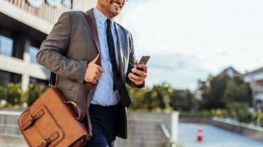 İyi giyimli profesyonel bir adam, güneşli bir günde dışarıda akıllı telefonunu kontrol ederken deri bir çantayla dolaşır..