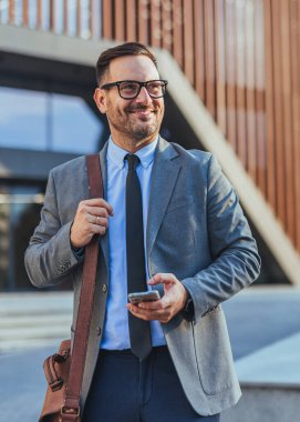 Kendine güvenen iş adamı takım elbise giyiyor, elinde akıllı bir telefon, modern bir ofis binasının önünde duruyor..