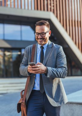 Takım elbiseli kendine güvenen bir profesyonel, şehir ortamında başarı, bağlantı ve profesyonelliği yansıtan çağdaş bir ofis binasının yakınındaki akıllı telefonunu kontrol ederken gülümsüyor..