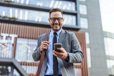 Modern bir binanın önünde dikilirken gülümseyen iş adamı omuzunda bir torbayla şık kıyafetler giyer iş günü boyunca güven ve bağlantı yayar..