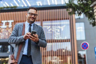 Takım elbiseli, açık havada akıllı telefon kullanan kendine güvenen işadamı, modern cam bir binanın yanında duruyor. Kurumsal bir ortamda başarı, iletişim, profesyonellik ve kentsel yaşam tarzını temsil ediyor..