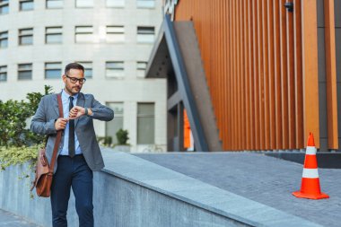 Kendine güveni tam bir işadamı, modern mimarinin yanında yürürken saatine bakıyor. Zamanlamayı, profesyonelliği ve şehir hayatı tarzını temsil eder.