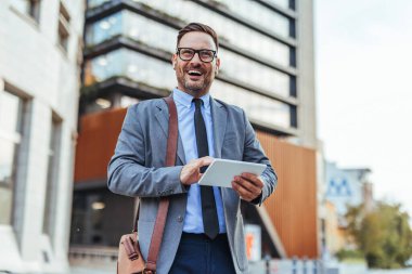Tablet kullanırken takım elbiseli başarılı bir işadamı güvenle gülümsüyor. Çağdaş mimarinin yanında yakalanan, profesyonellik, pozitiflik ve kentsel kurumsal bir ortamda başarı..