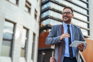 Resmi giyinmiş kendine güvenen bir işadamı tablet kullanırken gülümser, modern bir şehir merkezinde açık havada yakalanır. Görüntü profesyonelliği, başarıyı, güveni ve teknolojik katılımı aktarıyor.