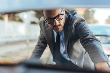 Takım elbiseli ve gözlüklü odaklanmış işadamı dışarıdaki araba motoru kompartımanını inceliyor. Güneşli bir günde resmedilen profesyonel ve ciddi davranış.