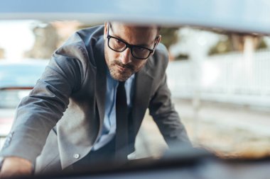 Gözlük takan profesyonel bir işadamı, bir arabanın bagajını kontrol eder. Açık hava ortamında, kendine güveni ve dikkati tam olur..