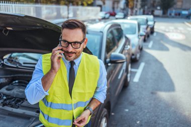 Yansıtıcı güvenlik yeleği giyen bir adam yol kenarında yardım isteyen açık kapüşonlu park halindeki bir arabanın yanından telefon eder. Şehir sokaklarında sakin ve sakin görünüyor..